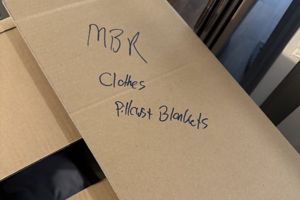 A moving box labeled by room in marker: master bedroom, clothes, pillows and blankets
