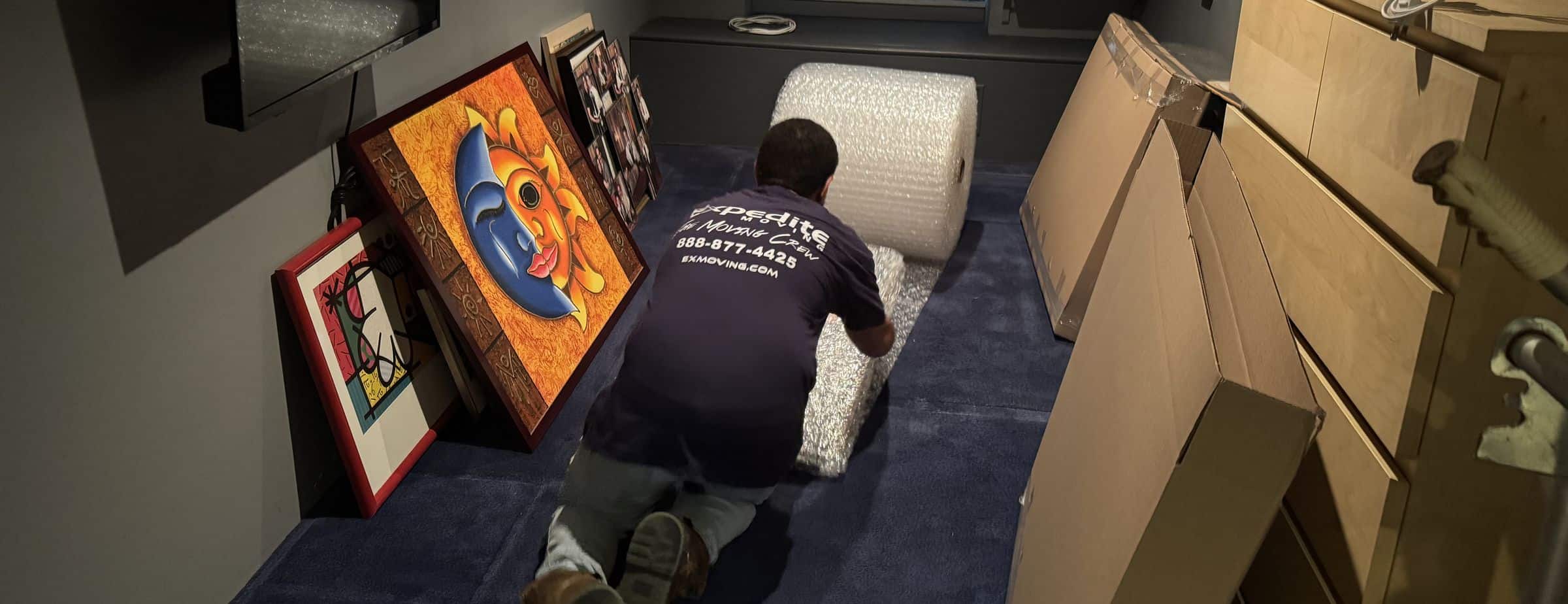 Expedite Moving mover unrolling bubble wrap to pack framed artwork beside stacked moving boxes in a New Jersey home
