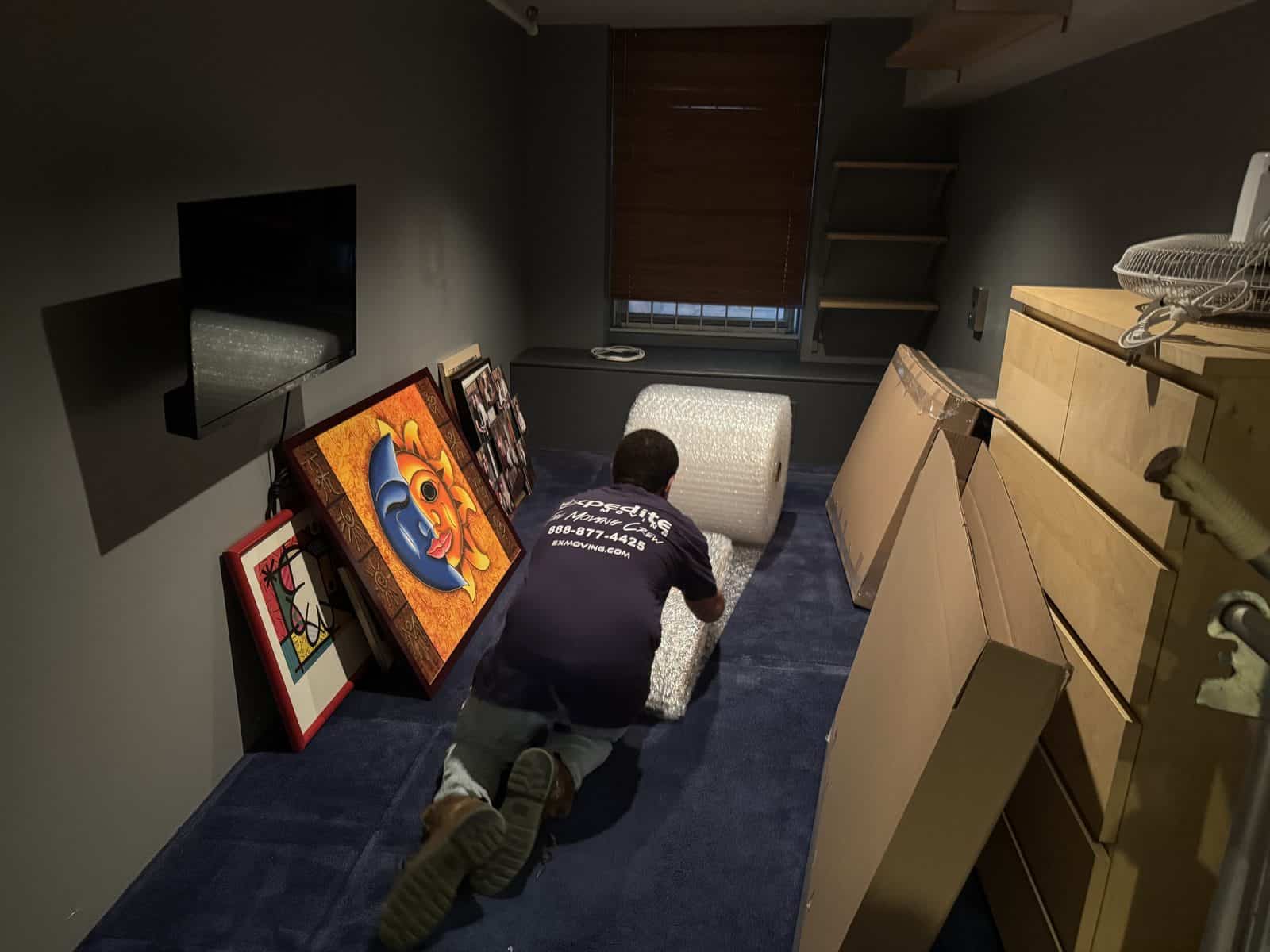 An Expedite Moving crew member unrolling bubble wrap to pack framed artwork in a New Jersey home, with flat picture cartons standing ready beside him